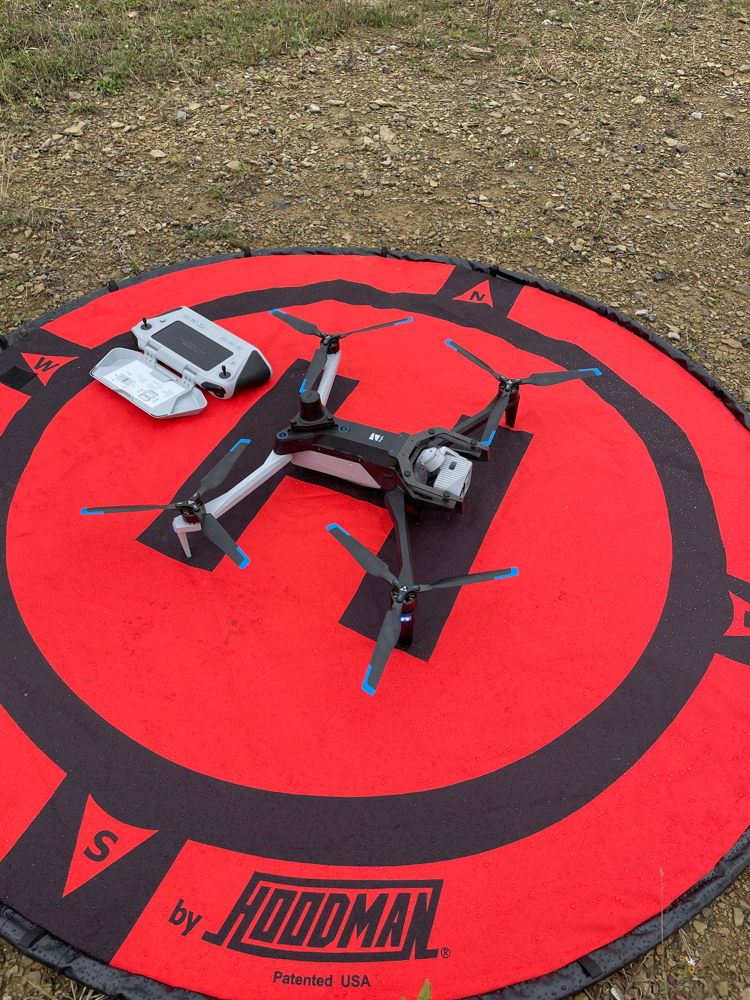 A 4 rotor copter style drone centered on it's circular launch pad. The dual stick control device is opened and laying on the launchpad behind the drone.
