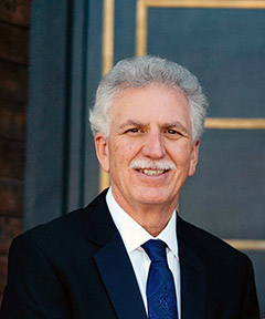 A headshot of Bill Jospeh, with gray hair and mustache, black suit and blue tie
