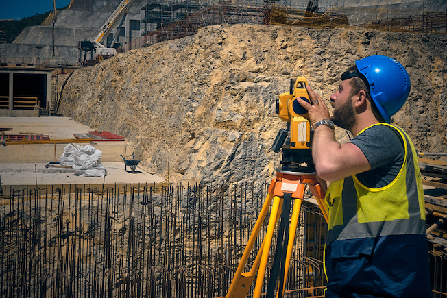image of Land surveyor stands by theodolite on a tripod against construction site at background. 