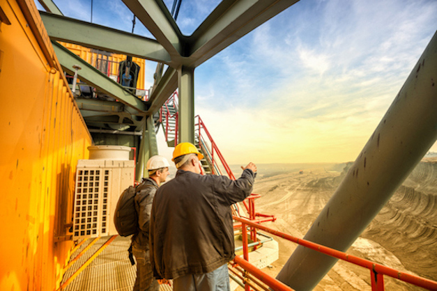 Image of Two coal mine engineers with protective helmets standing on a huge drill machine, chatting and watching at the digging site. Beautiful and colorful sky in the background. Rear view.