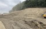 Three pieces of yellow heavy equipment stabilize a soil covered, gently sloping hillside below a treeline